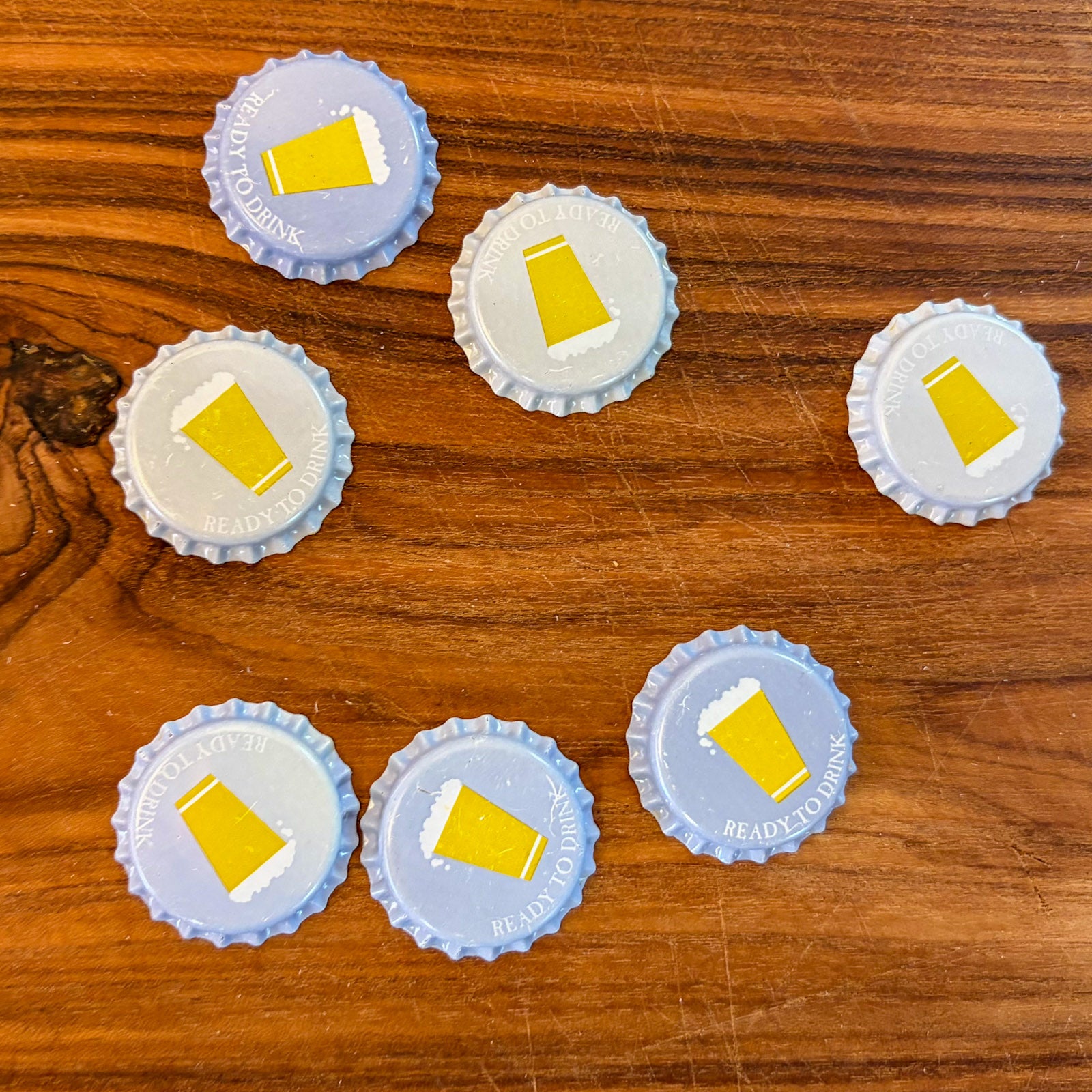 Six bottle caps with a beer pint illustration arranged on a wooden surface, three are slightly colored light blue to demonstrate cold activation, the other three are white