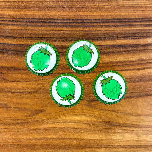 Four bottle caps with green hop illustrations on a white background, arranged on a brown wood surface.