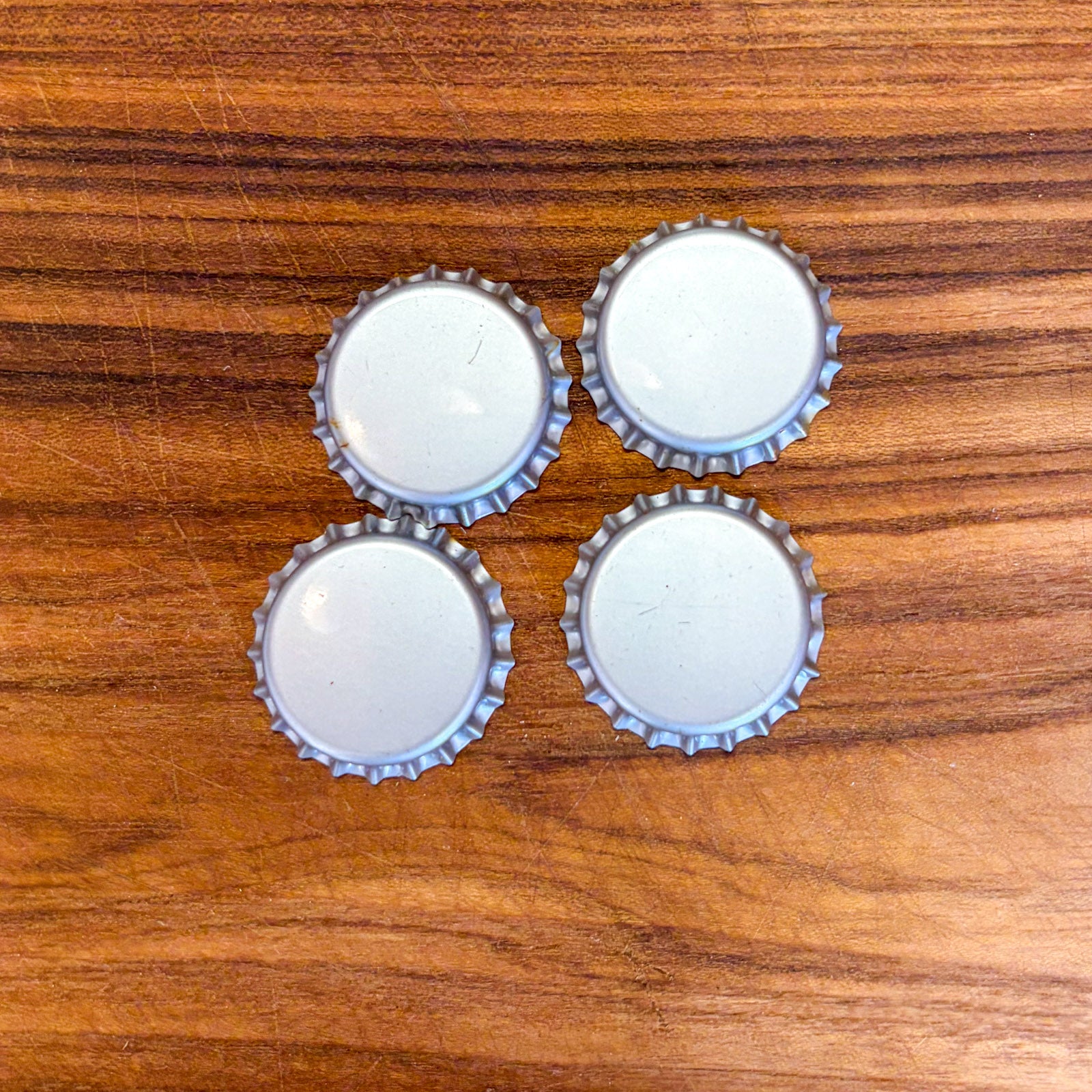 Four silver metal bottle cap covers arranged in a 2x2 grid on a brown wooden surface.