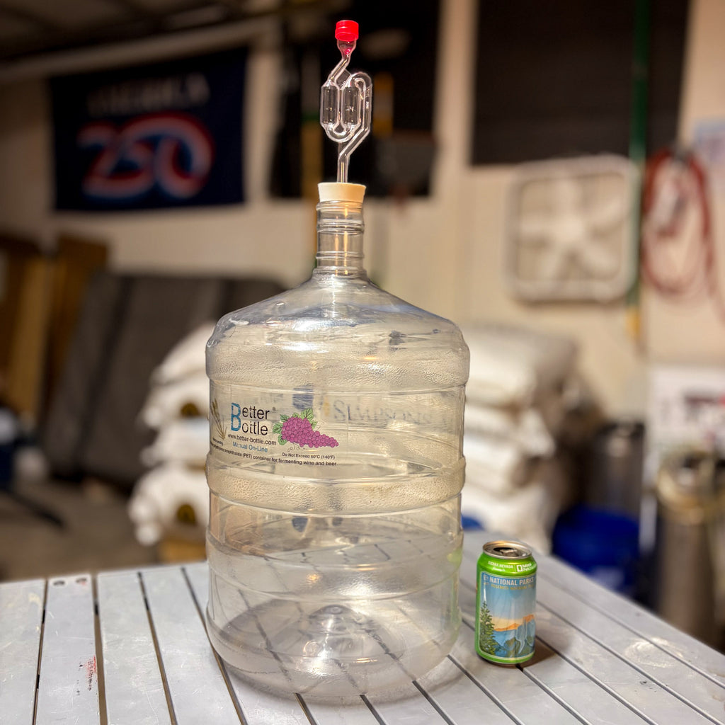 A 6 gallon clear and empty better bottle plastic carboy on a table with carboy stopper #10 and an s airlock attached