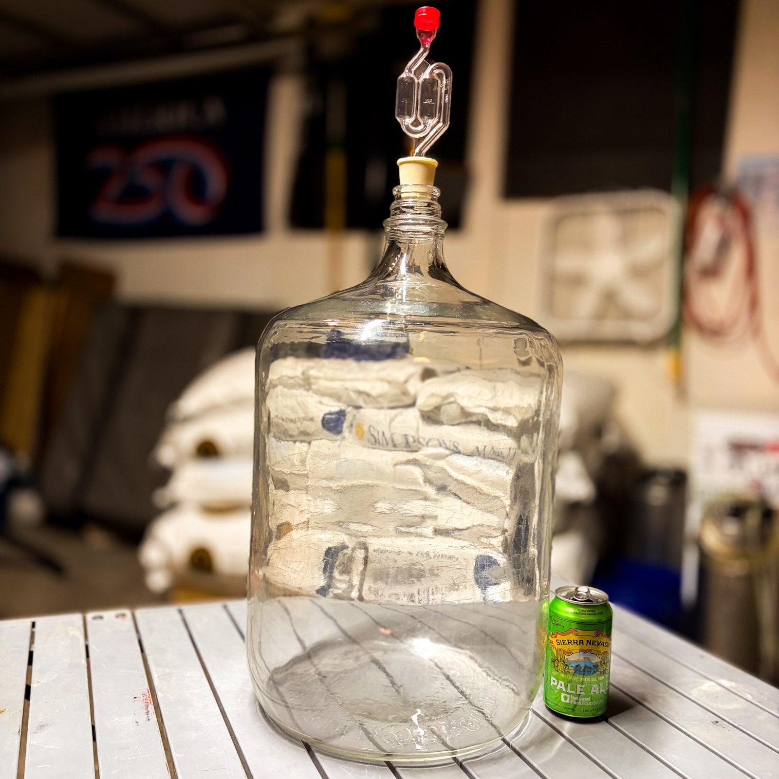 A carboy, bung and airlock on a table, with a grain sack background and a can of Sierra Nevada pale ale.