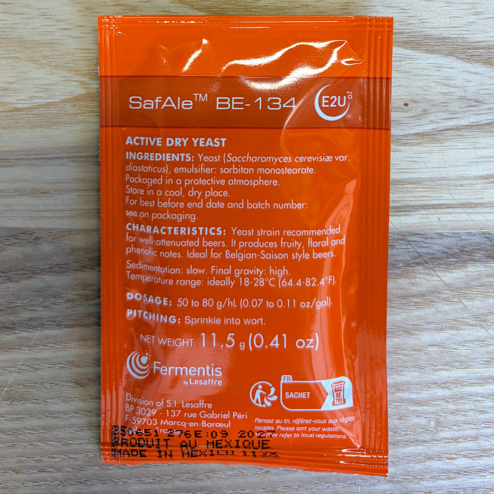 An orange fermentis SafAle BE-134 dry yeast sachet with white text, photographed flat on a light wood grain surface.
