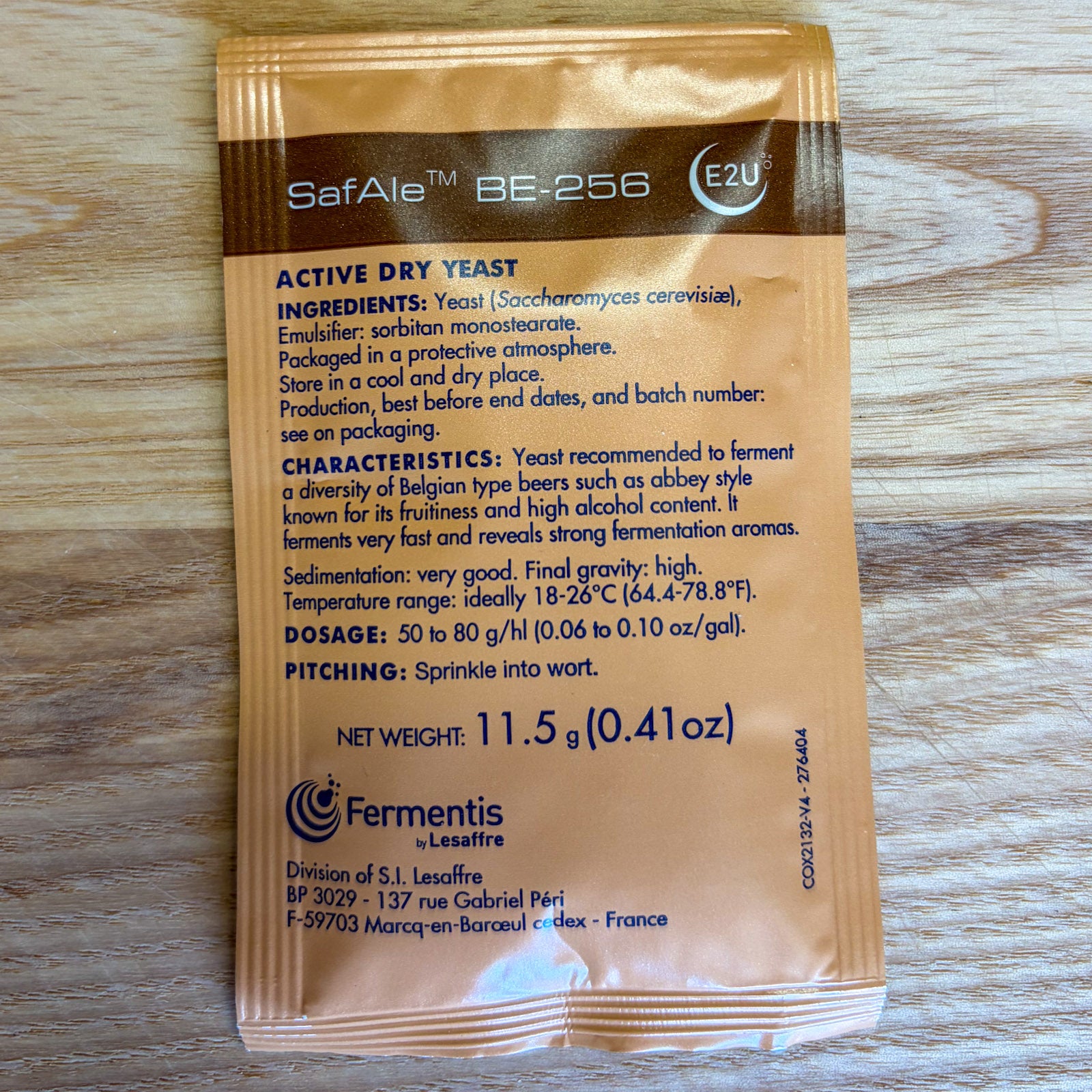 A single packet of SafAle BE-256 active dry yeast by Fermentis Lesaffre, shown flat on a light wood background. The packet is brown with black text detailing the product's ingredients, characteristics, dosage, and pitching instructions.