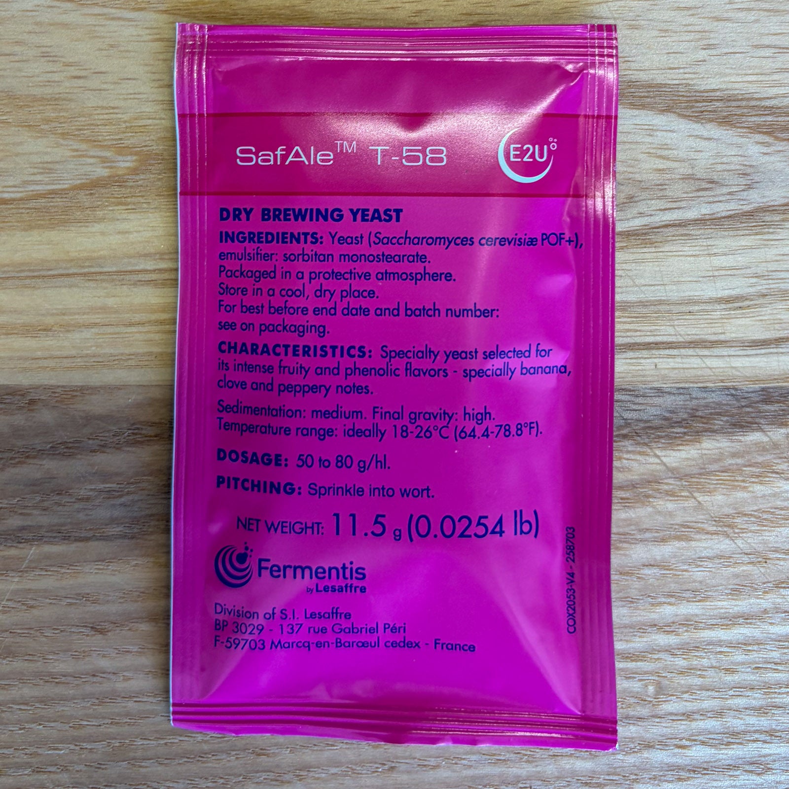 A single pink fermentis SafAle T-58 dry brewing yeast packet by Fermentis lies flat on a light wood surface. The front of the packet is printed with product details in white and light blue text.