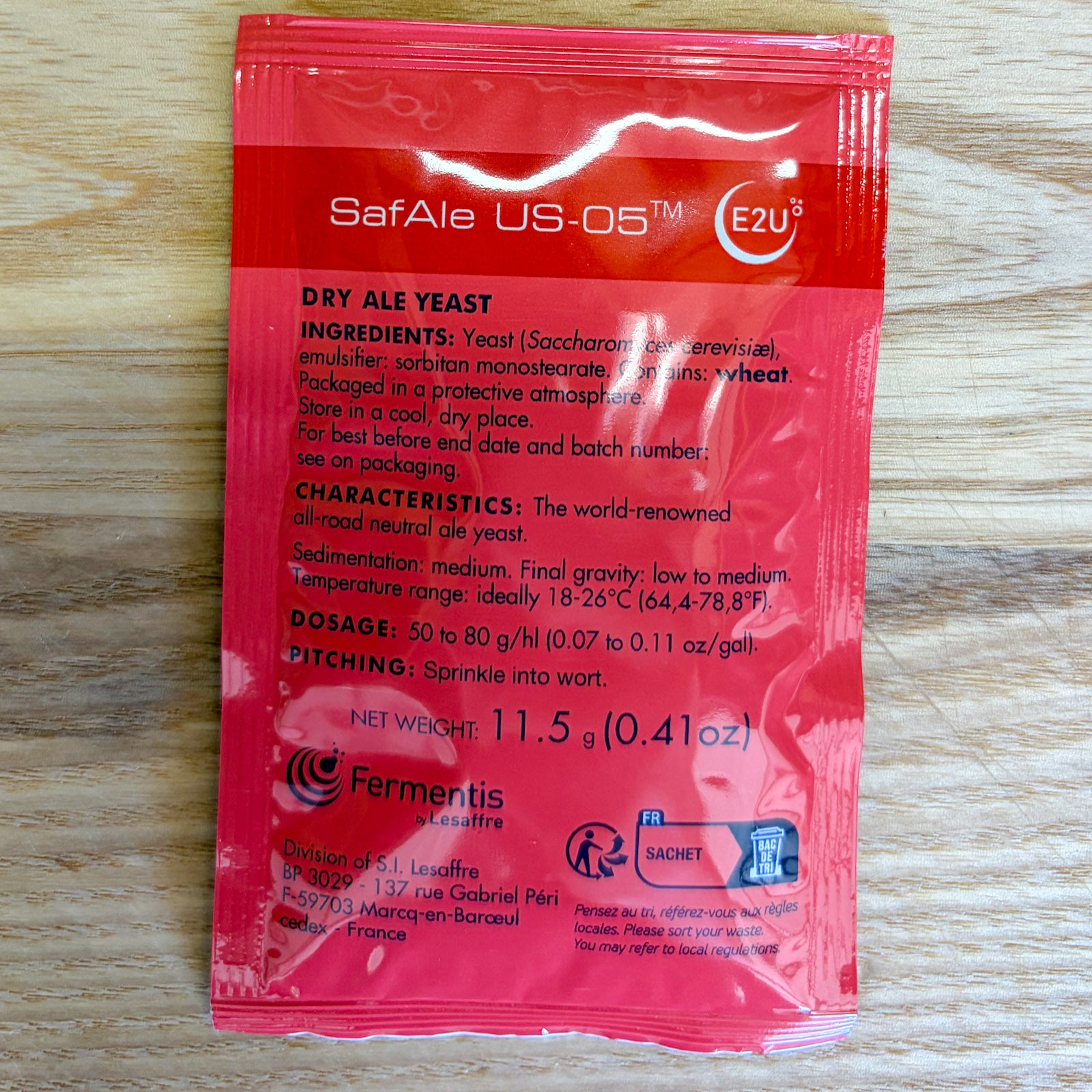 A red SafAle US-05m dry ale yeast packet by Fermentis s.A. Lesaffre, displayed flat on a light wood background.