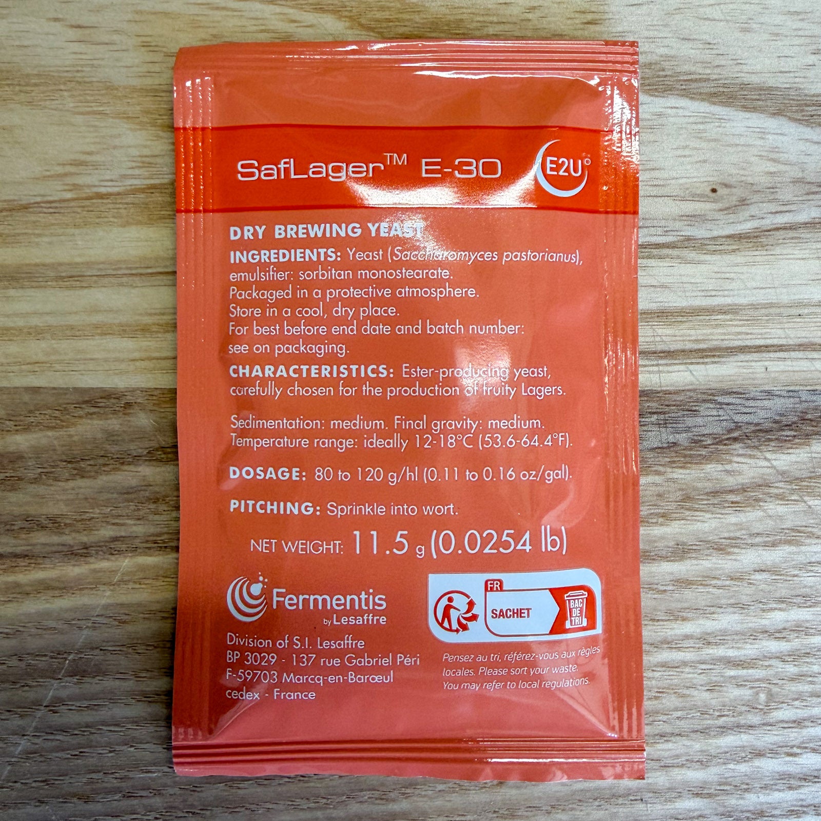 A single orange packet of Saflegen E-30 Dry Brewing Yeast by Fermentis lesaffre, placed flat on a light-colored wooden surface. The packet contains product details in white text and features a white and red logo.