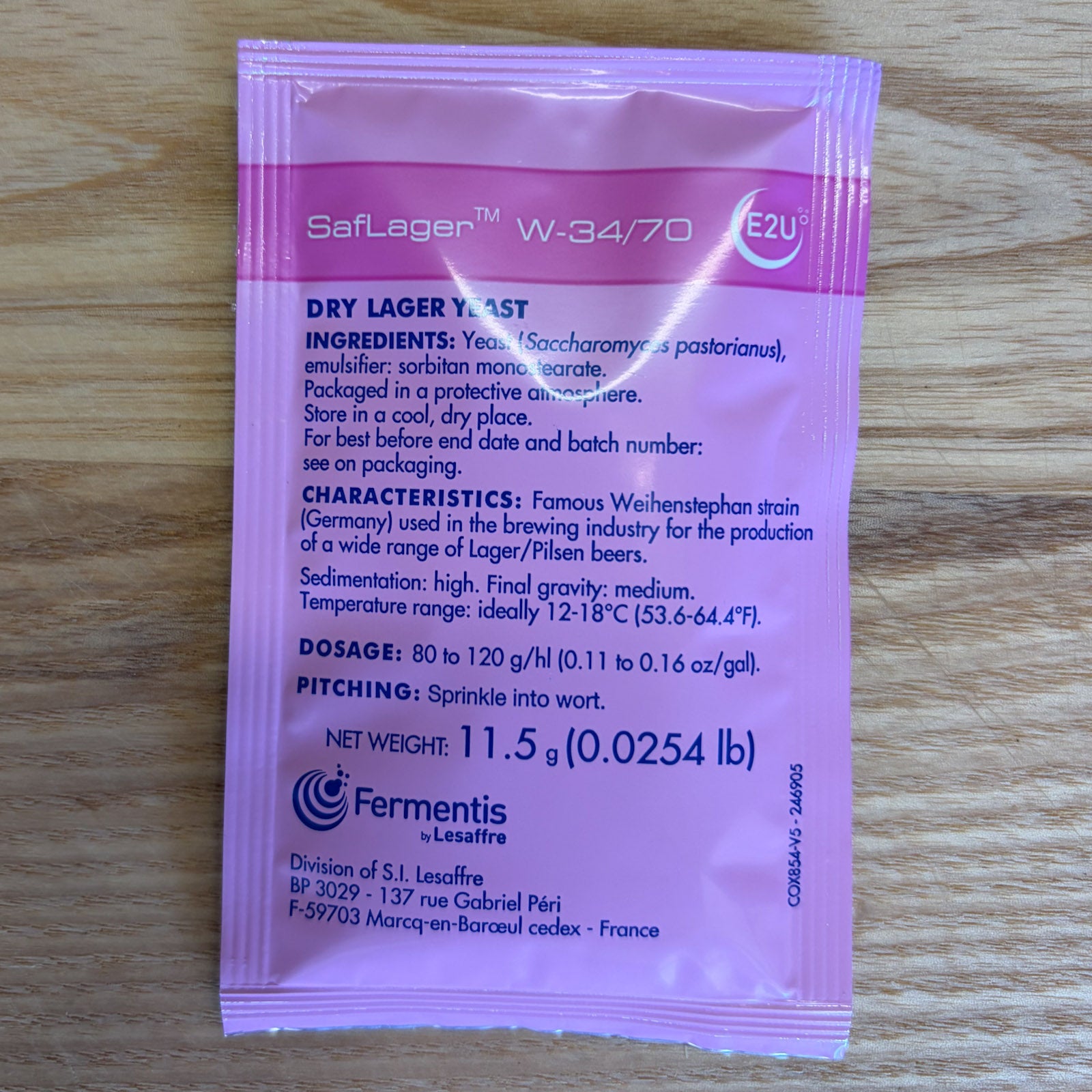 A single pink and white Fermentis SafLager W-34/70 dry lager yeast sachet lying flat on a light brown wooden surface. The front of the packet displays the product name, ingredients, characteristics, dosage, pitching instructions, and Fermentis branding.