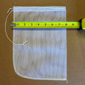 A square white mesh bag with a drawstring closure, shown on a brown cardboard background with a yellow tape measure laid across the top edge to indicate size.