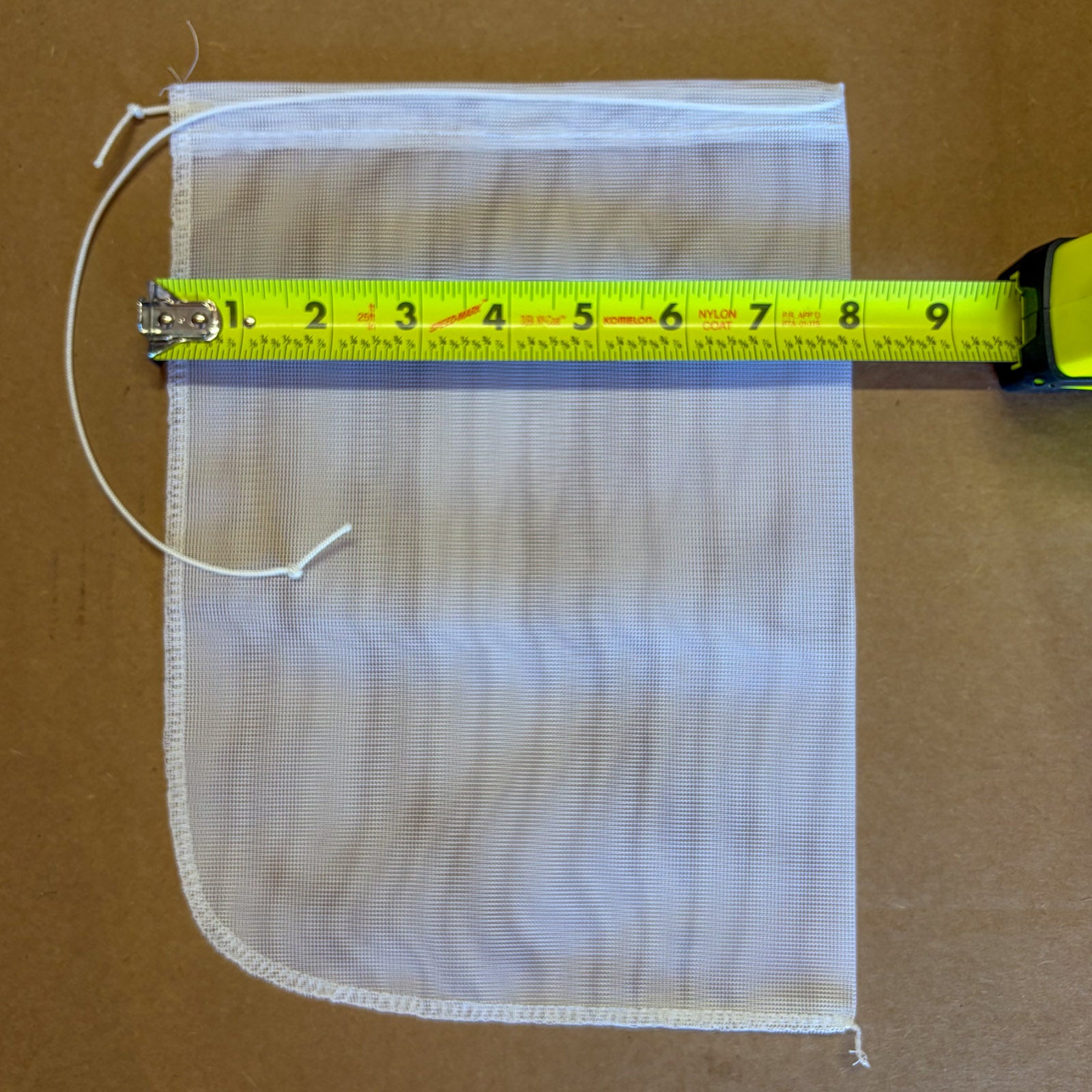 A square white mesh bag with a drawstring closure, shown on a brown cardboard background with a yellow tape measure laid across the top edge to indicate size.