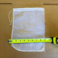 A white mesh drawstring bag laid flat on a brown cardboard surface, with a yellow tape measure stretched horizontally across the front to show its dimensions.