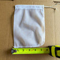 A white drawstring mesh bag laid flat on a brown cardboard surface, with a yellow tape measure placed along the bottom edge to show its size.
