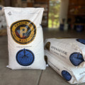 Two white bags of Proximity ReGenMalt base malted barley, with one standing upright and one lying down, on a stone floor in a rustic indoor setting.