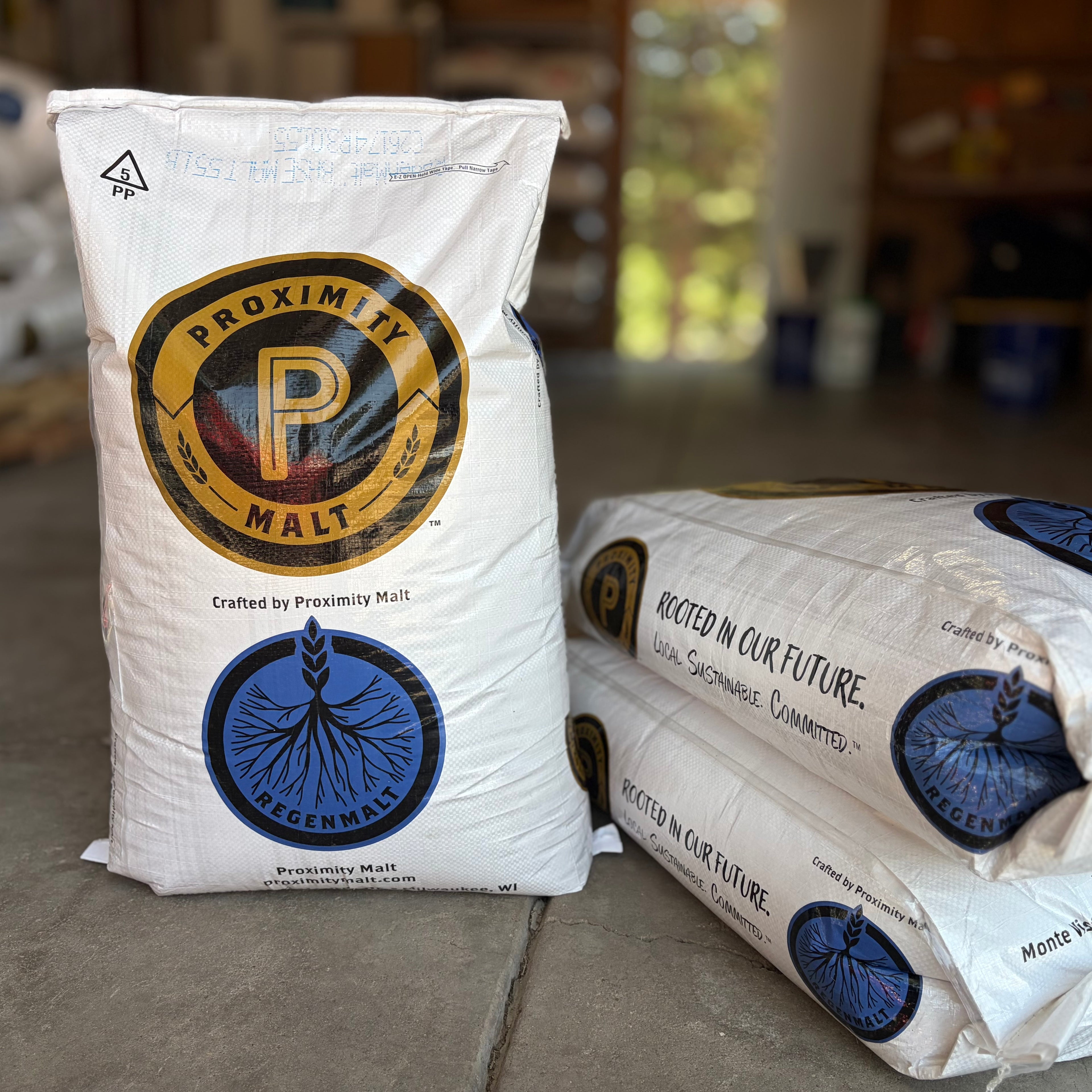 Two white bags of Proximity ReGenMalt base malted barley, with one standing upright and one lying down, on a stone floor in a rustic indoor setting.
