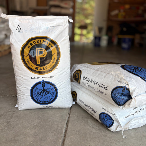 Two large white bags of Proximity regenMalt pale ale malt for brewing, with one standing upright and one lying on its side, placed on a concrete floor in a garage setting.