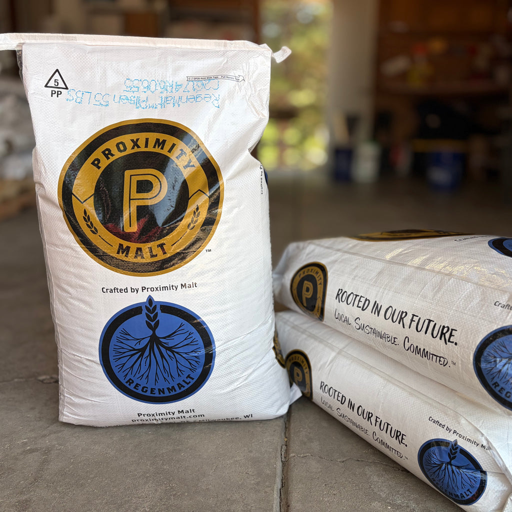 White bags of Proximity regenmalt pilsner malted barley, with one standing upright and two stacked on the floor in a garage setting