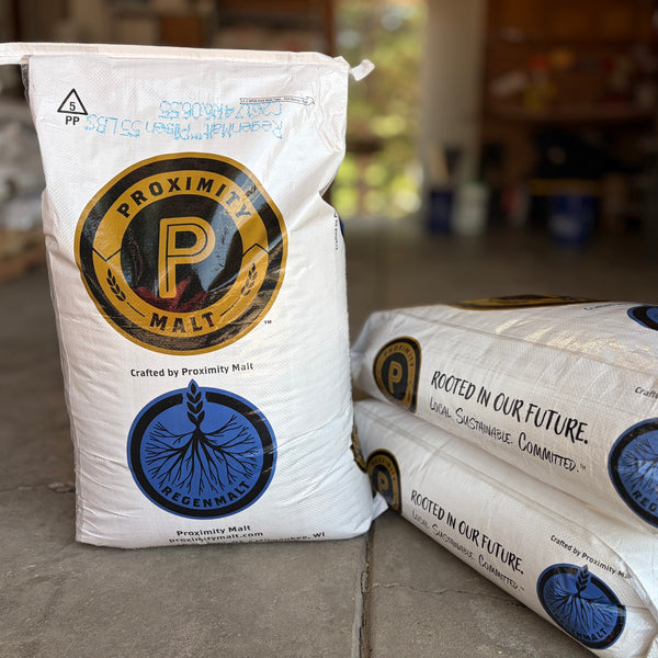 White bags of Proximity regenmalt pilsner malted barley, with one standing upright and two stacked on the floor in a garage setting