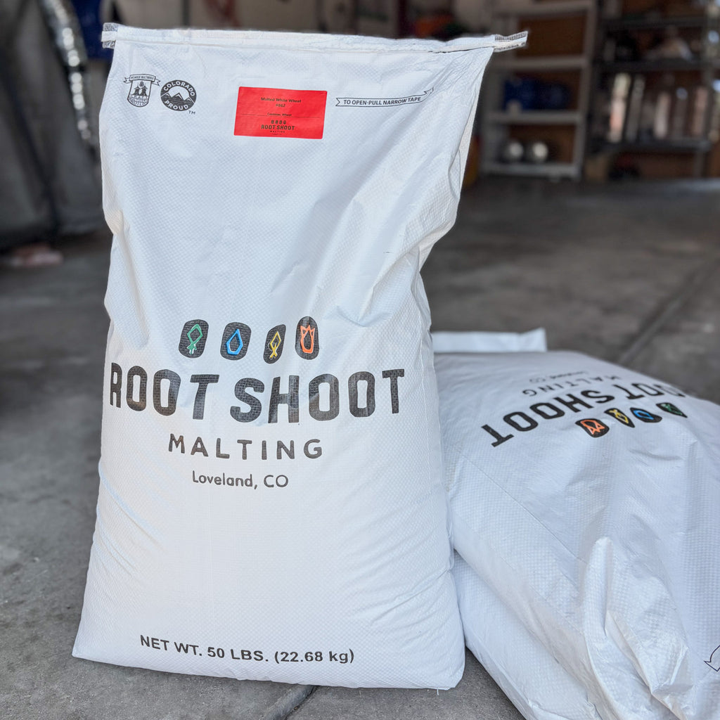A large white bag of Root Shoot Malting white wheat malt standing on a concrete floor, with a second bag partially visible behind it. The front bag shows the Root Shoot Malting logo, a red label, and text indicating Loveland, CO and net weight 50 lbs.