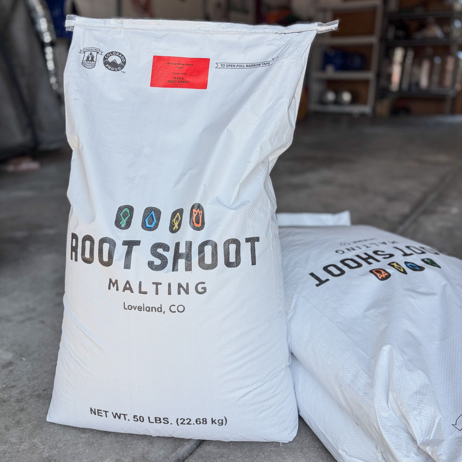A large white bag of Root Shoot Malting white wheat malt standing on a concrete floor, with a second bag partially visible behind it. The front bag shows the Root Shoot Malting logo, a red label, and text indicating Loveland, CO and net weight 50 lbs.