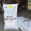 Two large white bags of Root Shoot Malting pilsner grain standing on a concrete floor in a garage-like setting. The front bag is upright and clearly shows the brand and product name, with a second bag lying beside it.