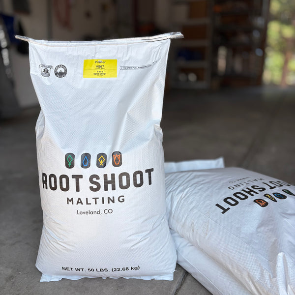 Two large white bags of Root Shoot Malting pilsner grain standing on a concrete floor in a garage-like setting. The front bag is upright and clearly shows the brand and product name, with a second bag lying beside it.
