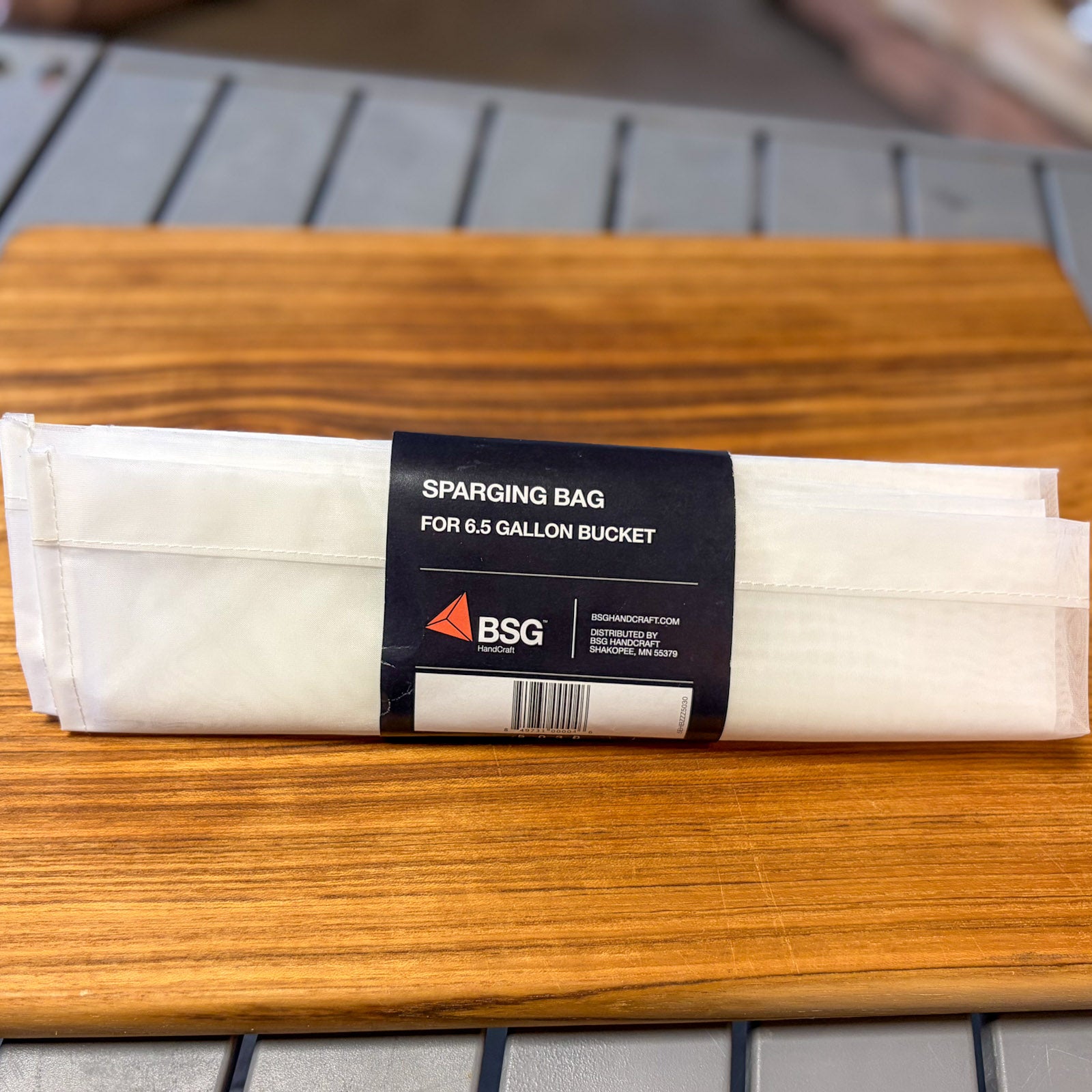 A packaged sparging bag for a 6.5 gallon bucket, wrapped in a black paper band with the BSG logo, resting on a wooden cutting board.