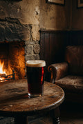 Glass of dark beer on a wooden table in an old cozy room with a fireplace and leather chair with fuggle hops