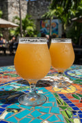 Two glasses of orange beer on a colorful mosaic table with a blurred tropical outdoor patio