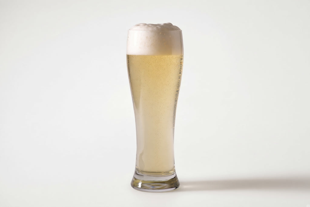A tall, slender clear glass filled with pale golden beer and topped with a thick white foam head, photographed on a clean light gray studio background with proximity regenmalt base malted barley