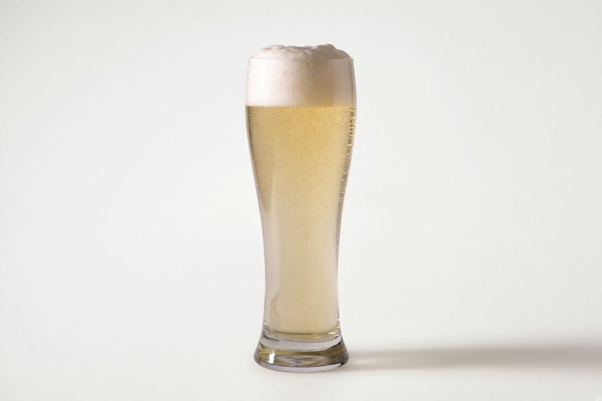 A tall, slender clear glass filled with pale golden beer and topped with a thick white foam head, photographed on a clean light gray studio background with proximity regenmalt base malted barley